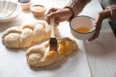 Photo of Woman spreading egg yolk over braided dough at white wooden table in kitchen, closeup. Cooking traditional Shabbat challah