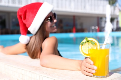 Photo of Young woman wearing Santa Claus hat with refreshing drink in swimming pool, focus on hand. Christmas vacation