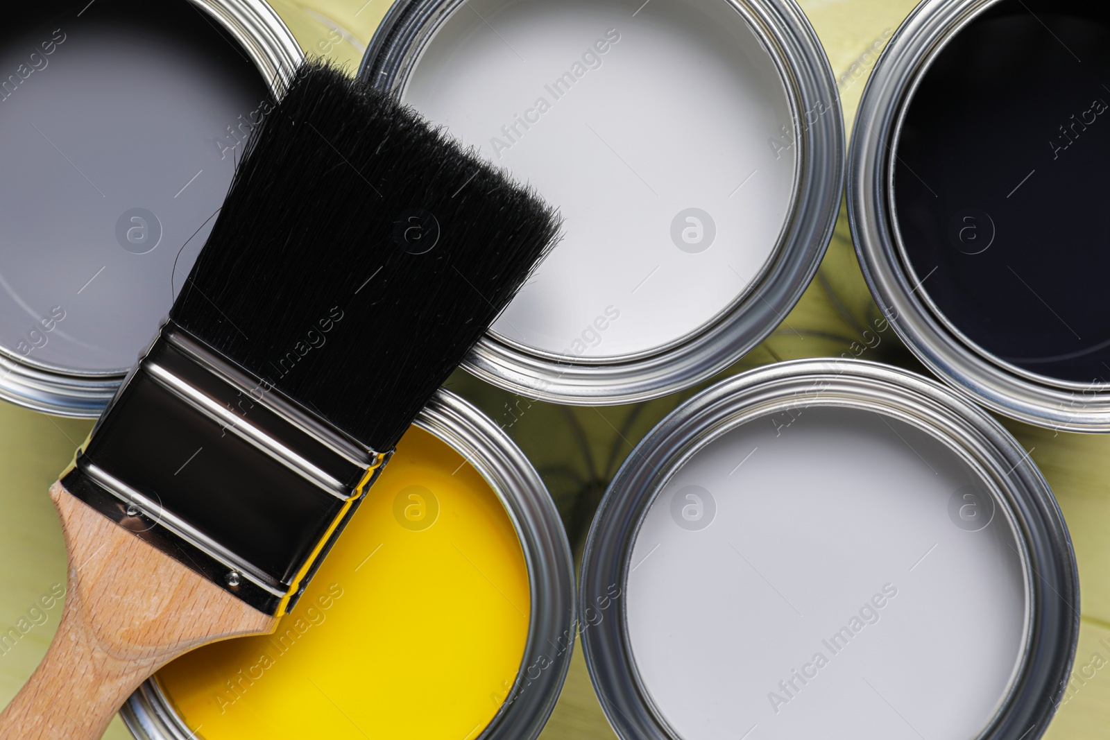 Photo of Cans of white, black, yellow and grey paints with brush on olive wooden table, flat lay