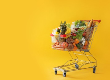 Photo of Shopping cart full of groceries on yellow background. Space for text