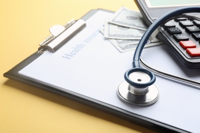 Photo of Clipboard with with medical insurance form, money, calculator and stethoscope on yellow background, closeup