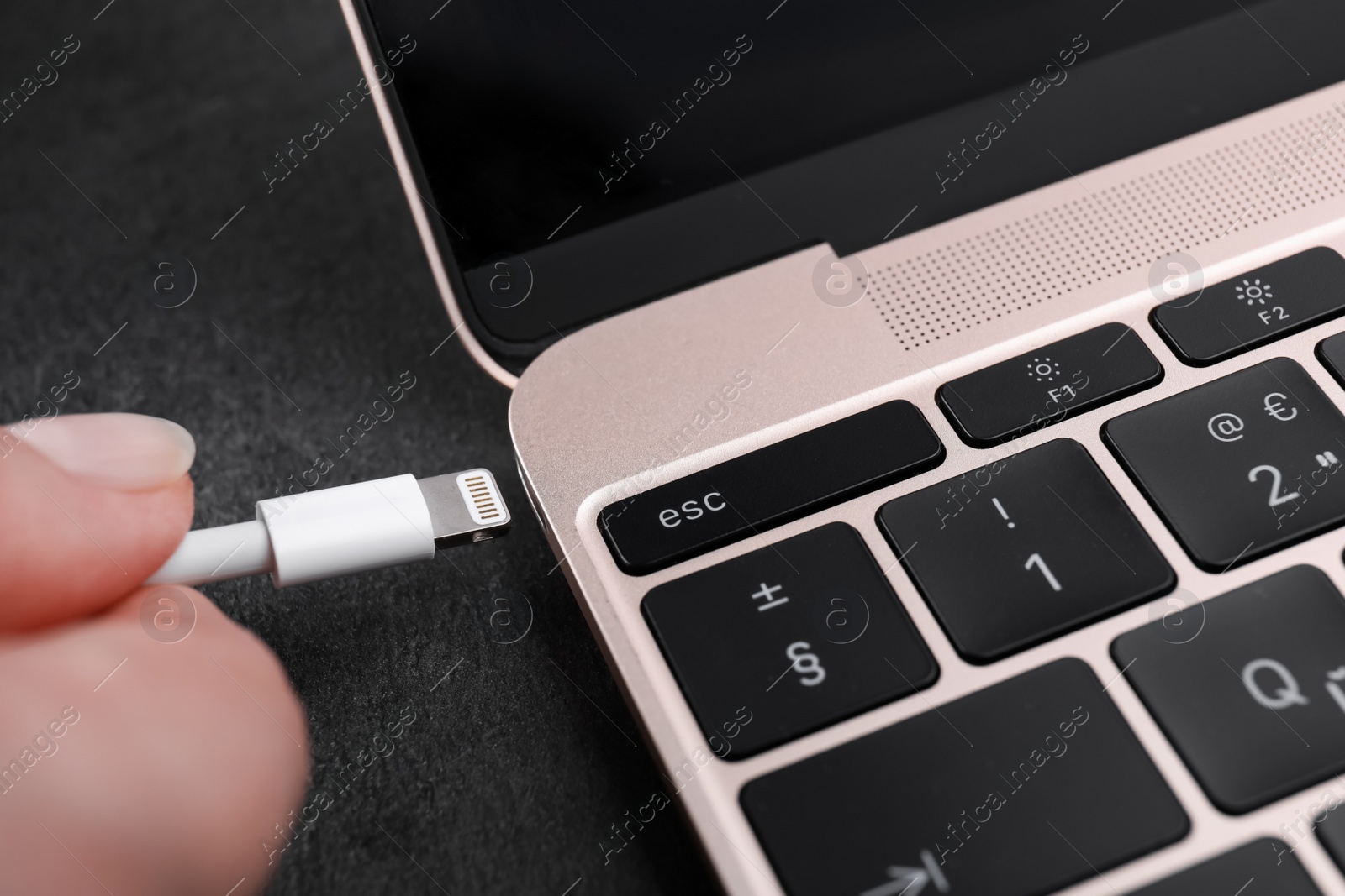 Photo of Woman plugging USB cable with lightning connector into laptop port on dark table, closeup