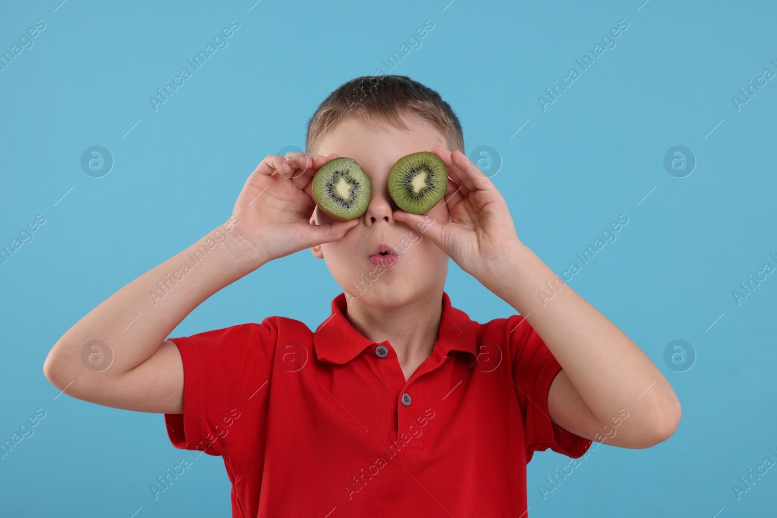 Photo of Emotional boy covering eyes with halves of fresh kiwi on light blue background