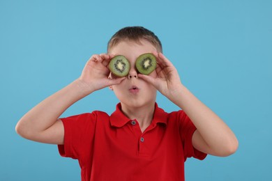 Photo of Emotional boy covering eyes with halves of fresh kiwi on light blue background