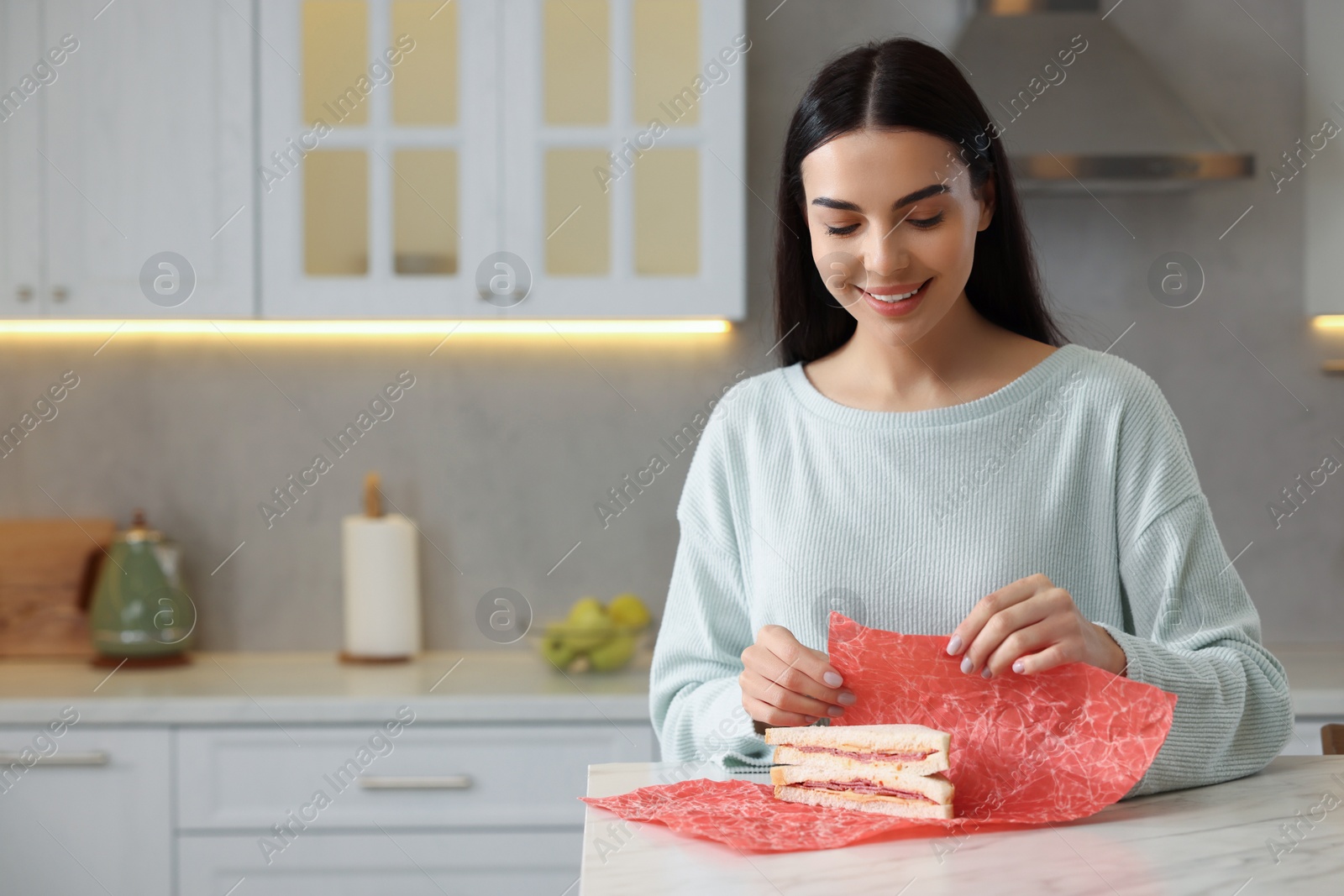 Photo of Happy woman packing sandwich into beeswax food wrap at table in kitchen