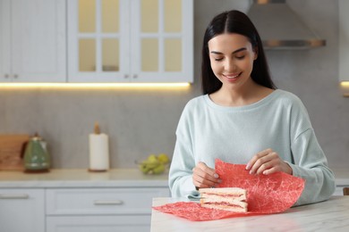 Happy woman packing sandwich into beeswax food wrap at table in kitchen