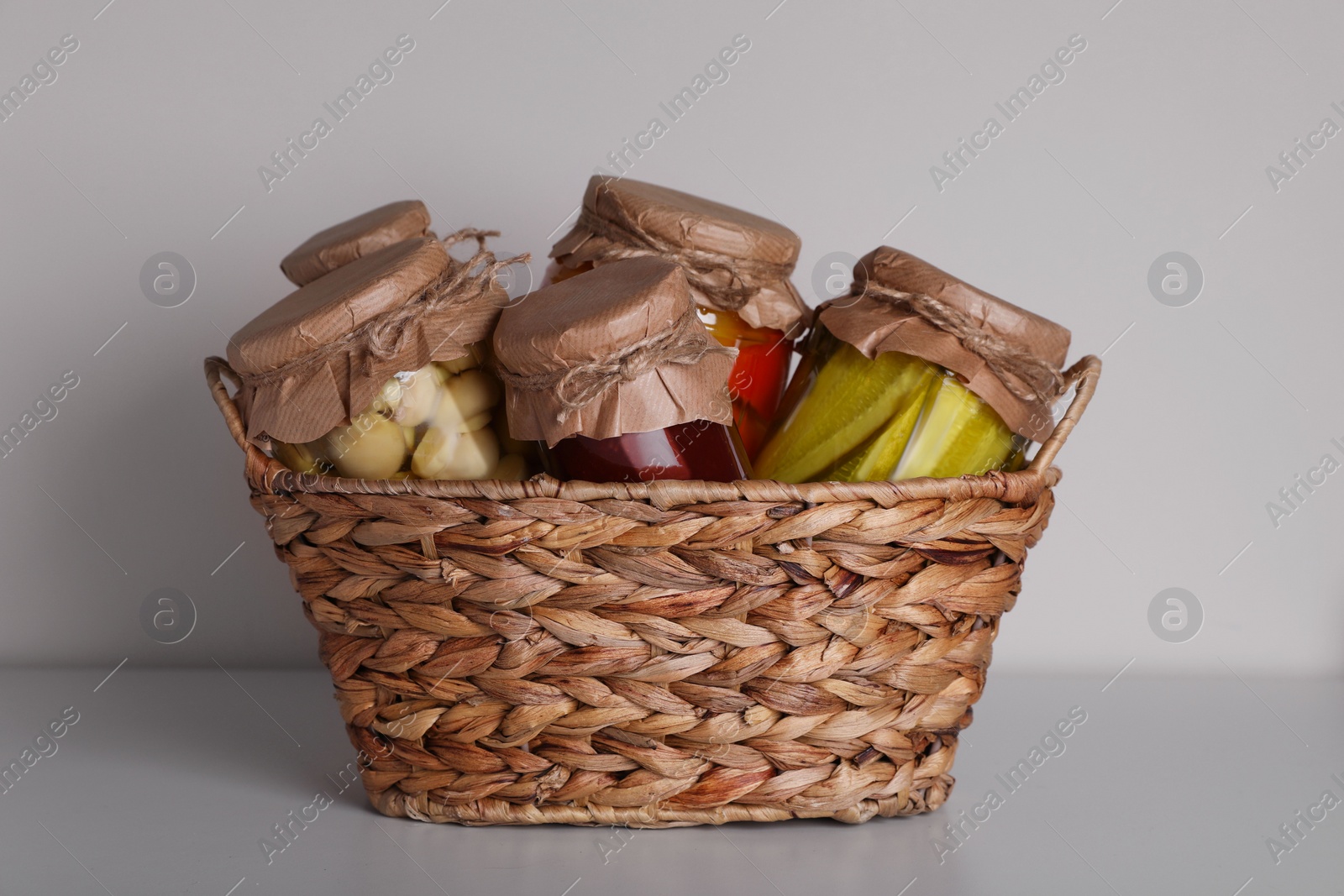 Photo of Wicker basket with many jars of different preserved products on light grey table