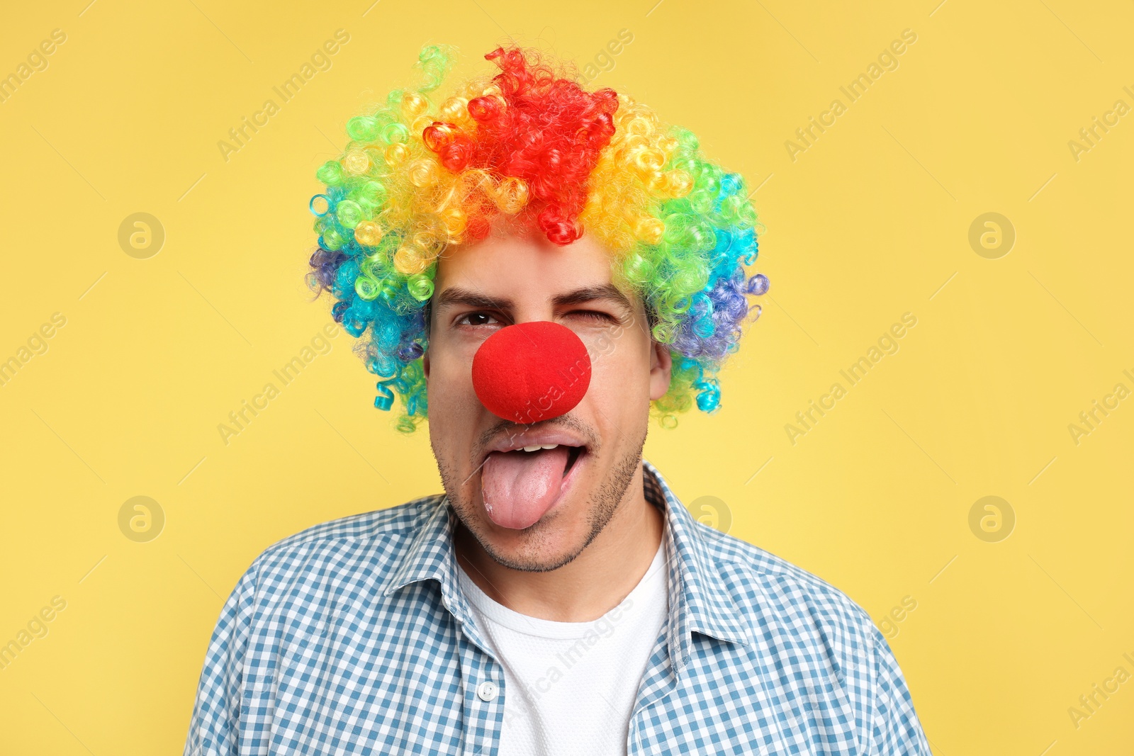 Photo of Funny man with clown nose and rainbow wig on yellow background. April fool's day