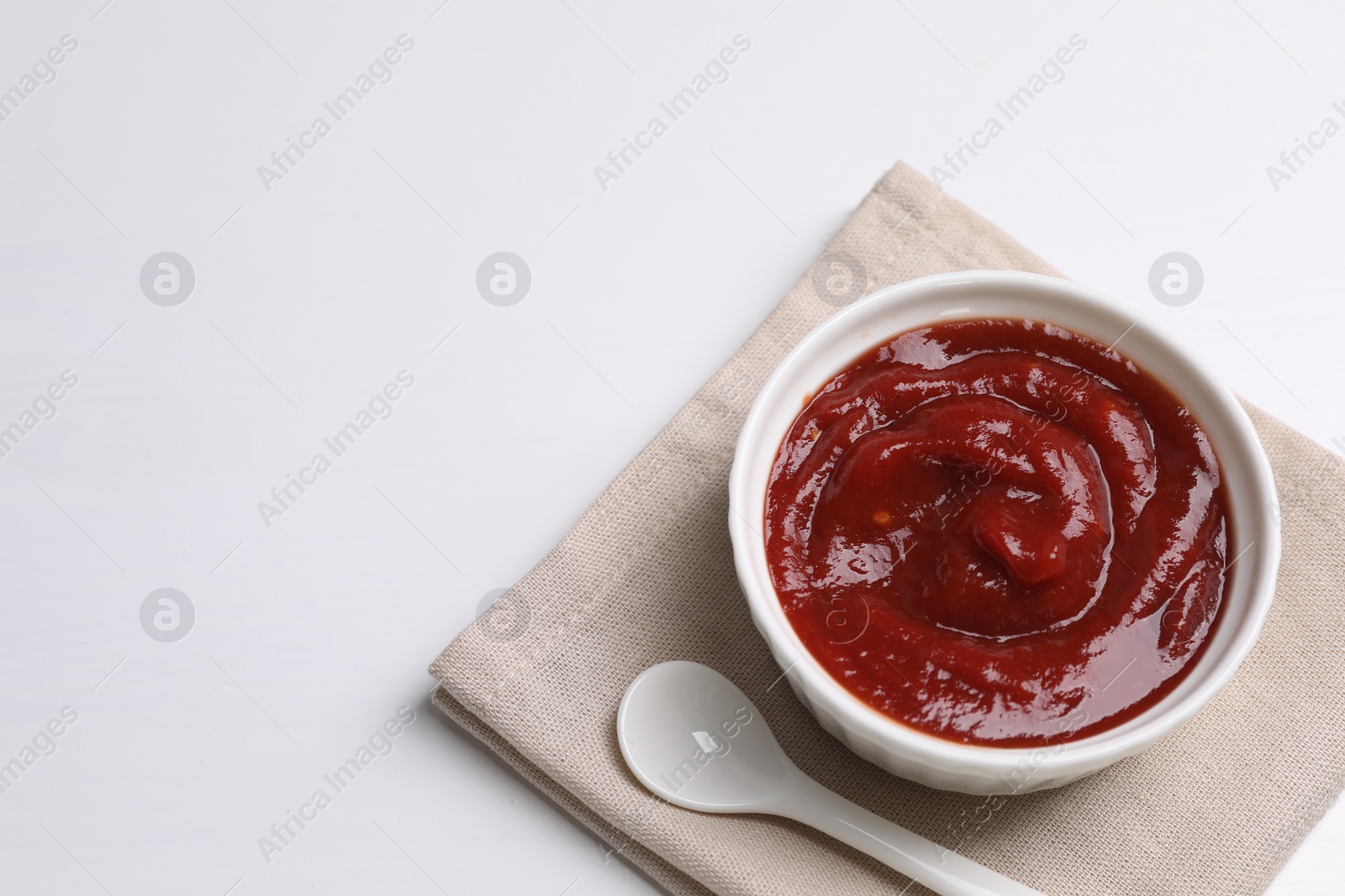 Photo of Organic ketchup in bowl and spoon on white table, space for text. Tomato sauce