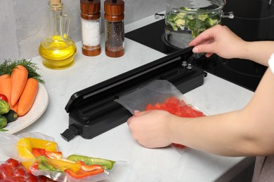 Photo of Woman using sealer for vacuum packing with plastic bag of red pepper at white table, closeup