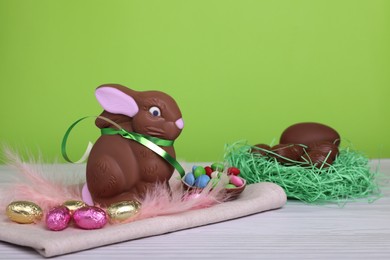 Photo of Chocolate Easter bunny, eggs, feathers and candies on white wooden table against light green background