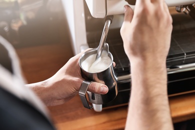 Photo of Barista frothing milk in metal pitcher with coffee machine wand at bar counter, closeup