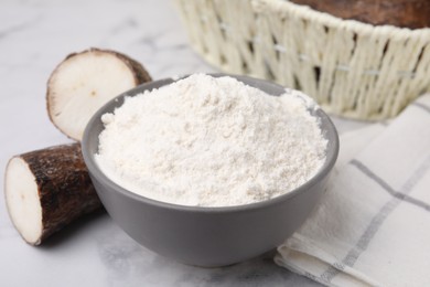 Photo of Bowl with cassava flour and roots on white marble table, closeup