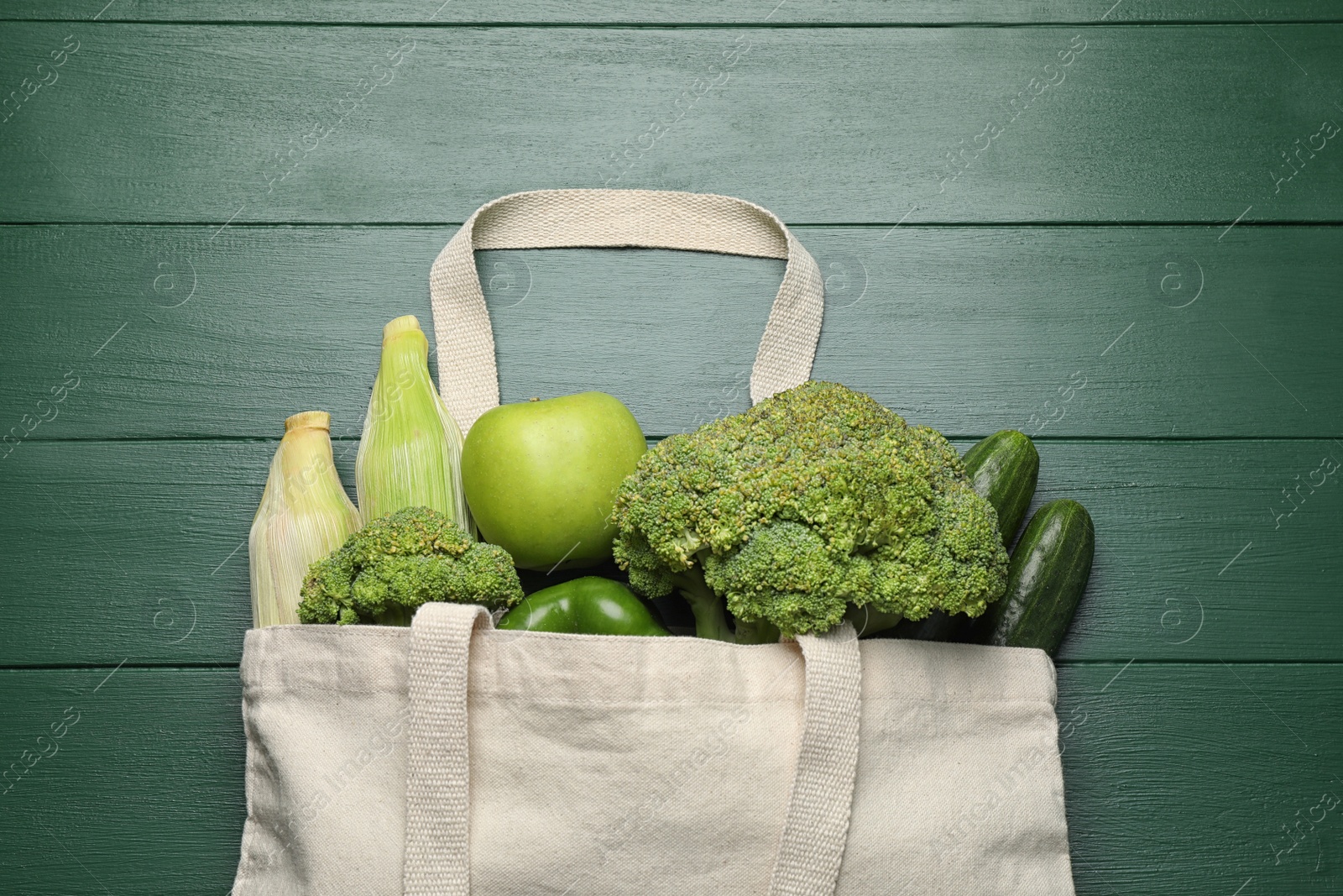 Photo of Cloth bag with fresh vegetables and apple on green wooden table, flat lay