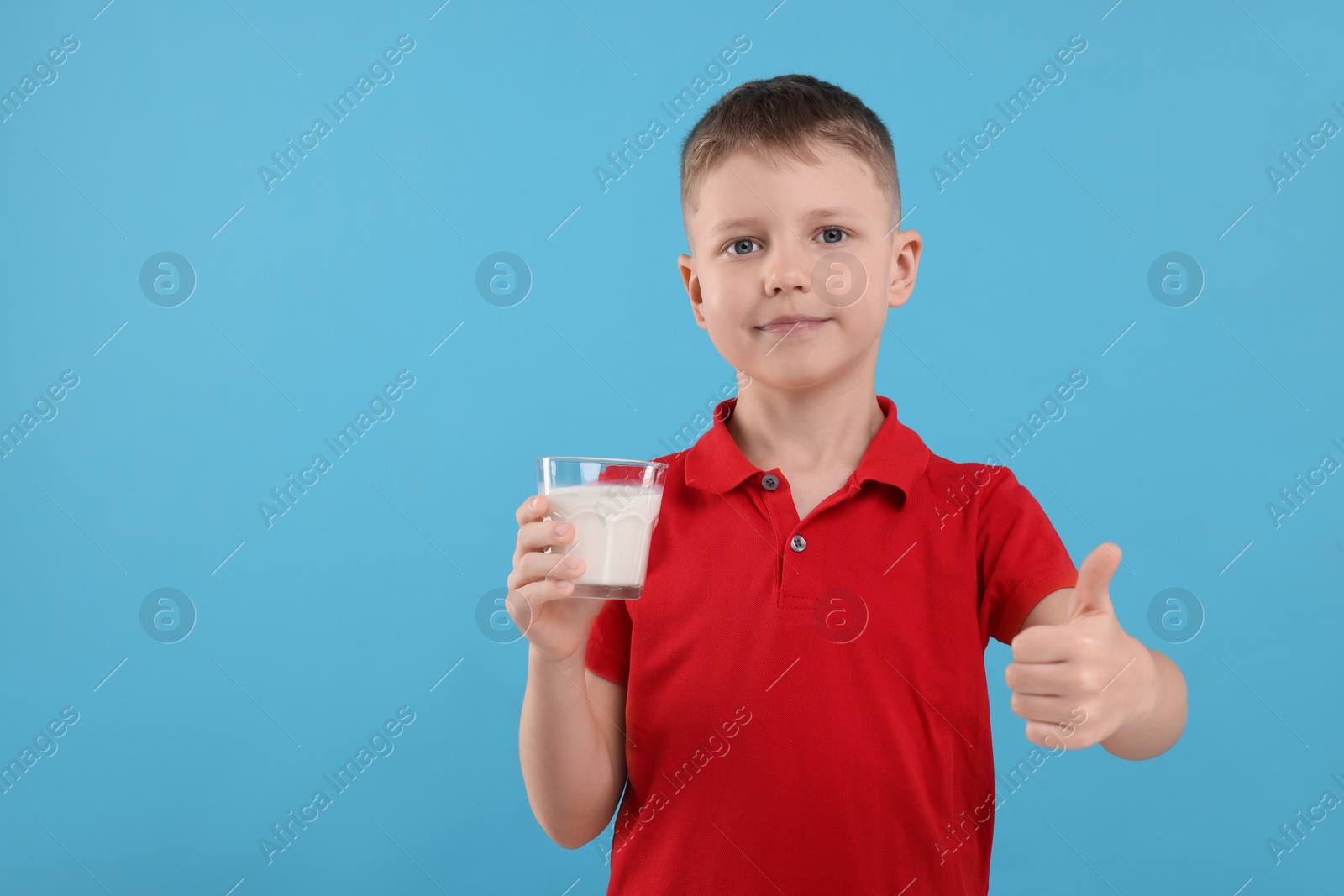 Photo of Cute boy with glass of fresh milk showing thumb up on light blue background, space for text