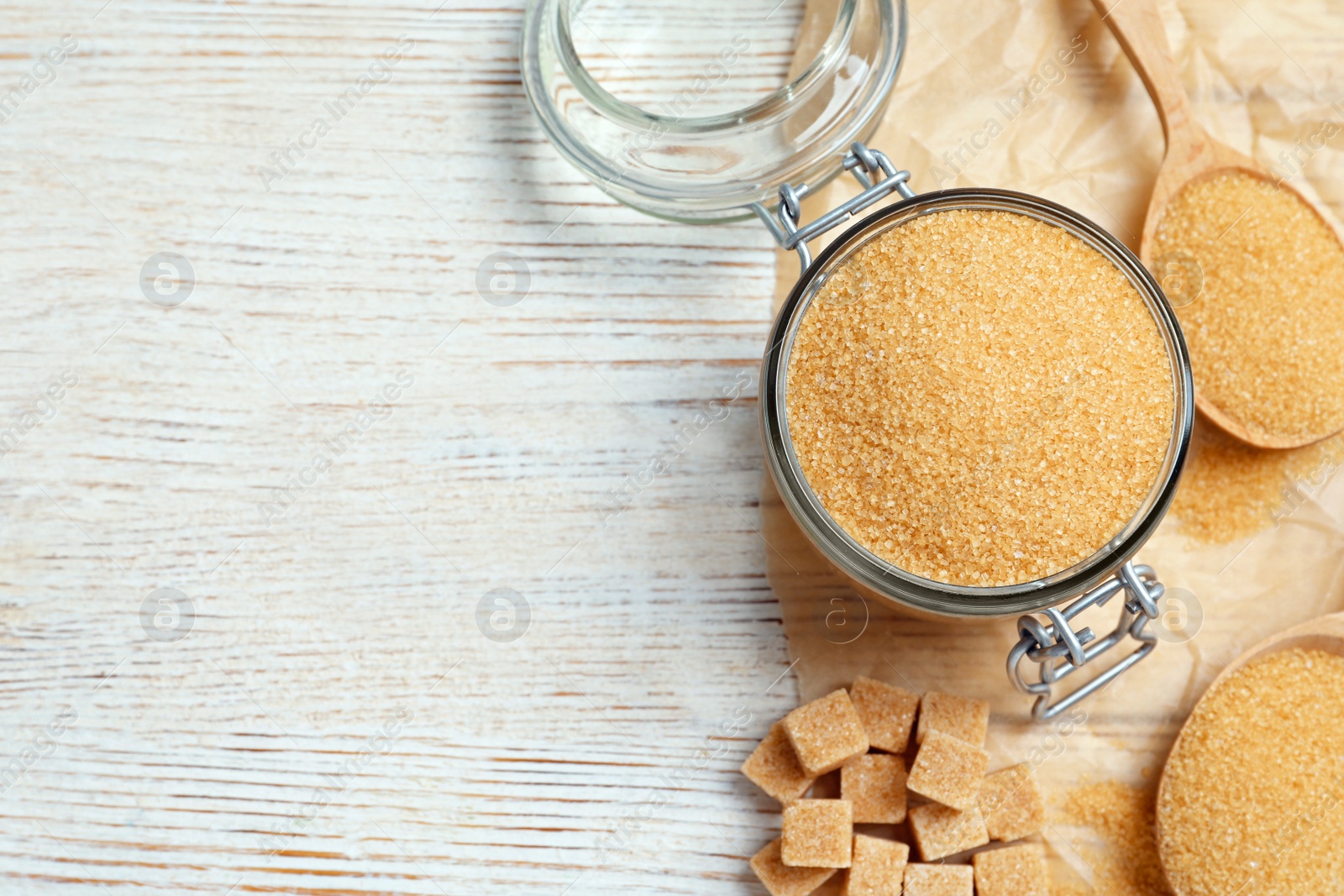 Photo of Glass jar of brown granulated sugar, spoon and cubes on light wooden table, flat lay. Space for text