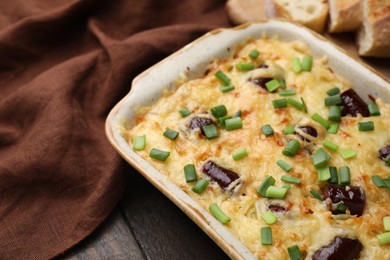 Photo of Tasty sausage casserole with green onions in baking dish on wooden table, closeup