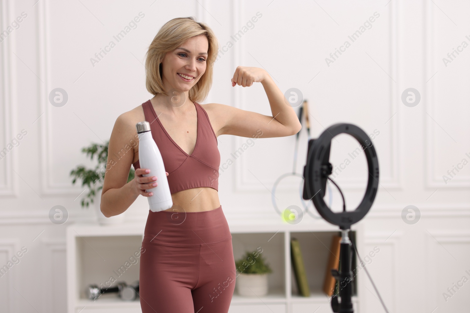 Photo of Smiling sports blogger holding bottle while streaming online fitness lesson at home