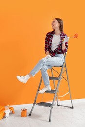 Photo of Designer with roller sitting on folding ladder near freshly painted orange wall indoors