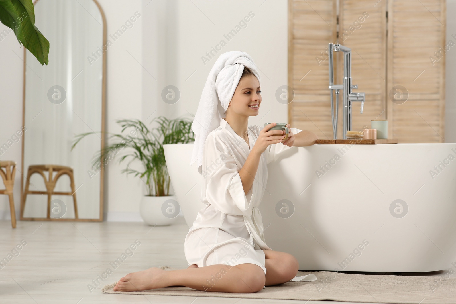 Photo of Beautiful happy woman with cup of hot drink in robe sitting on floor in bathroom