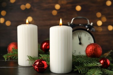 Photo of Burning candles, festive decor and alarm clock on wooden table against blurred Christmas lights