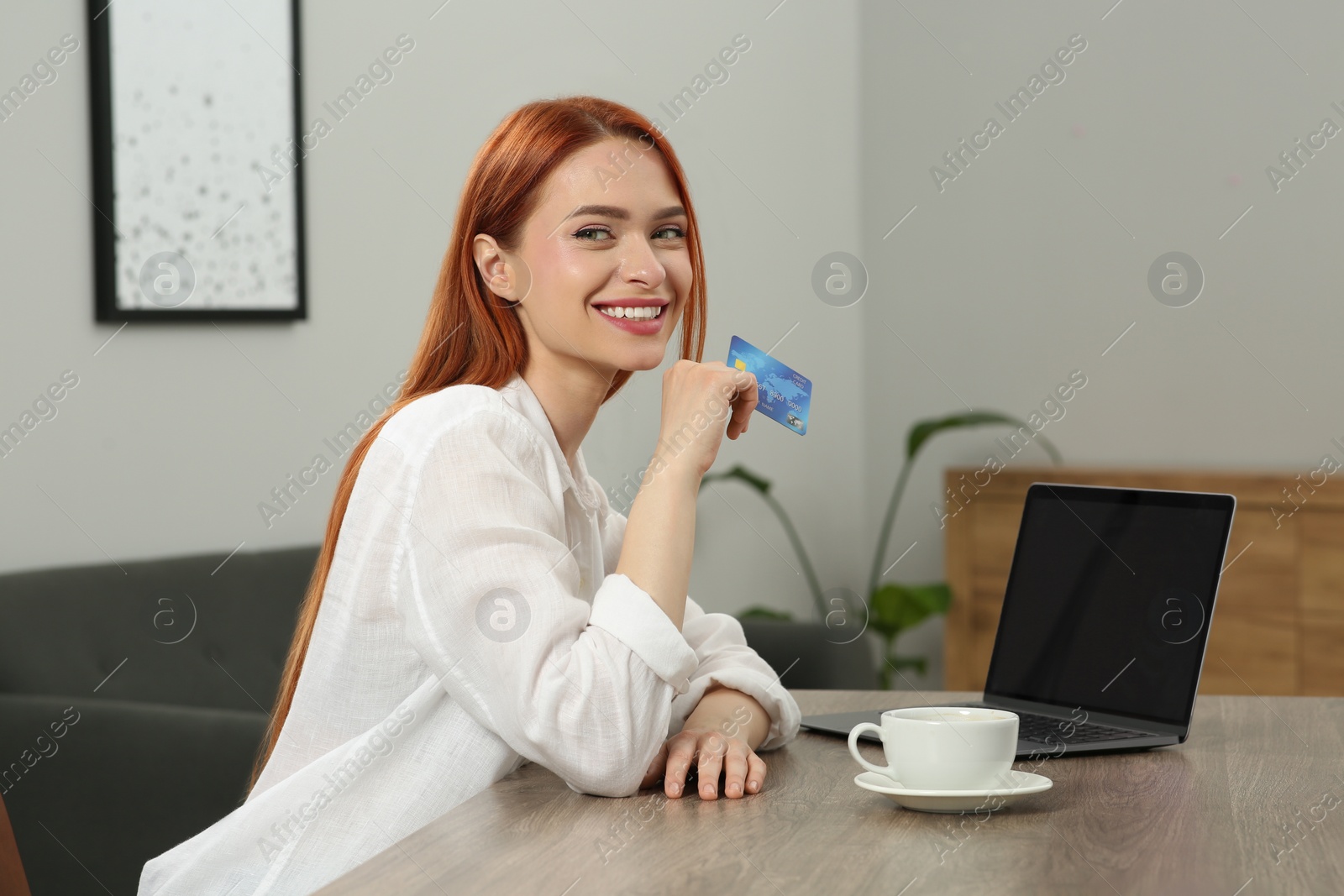 Photo of Happy woman with credit card near laptop at wooden table in room. Online shopping