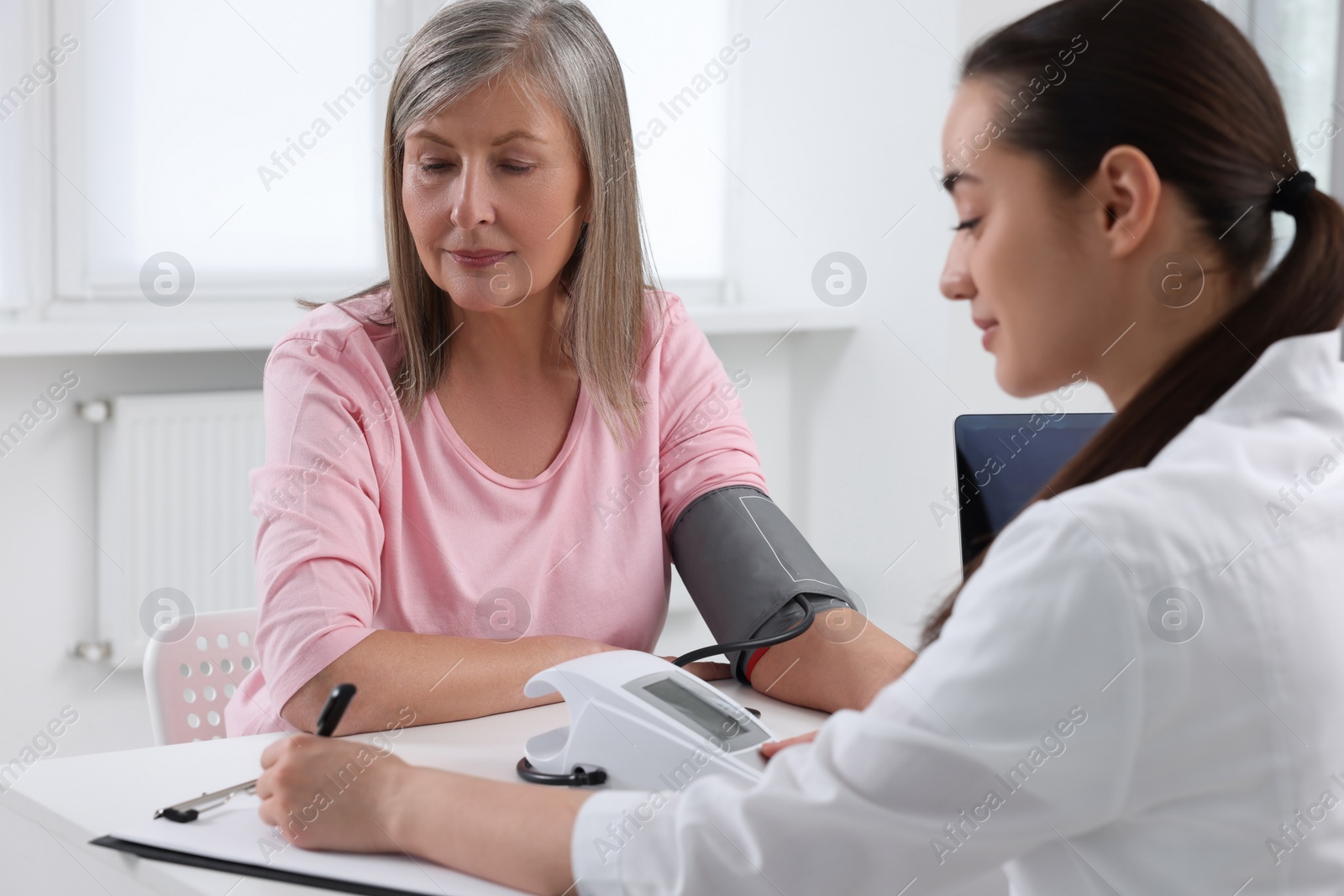 Photo of Doctor measuring blood pressure of woman and writing it down into clipboard at table indoors
