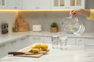 Photo of Woman pouring water from jug into glass at white table in kitchen, closeup