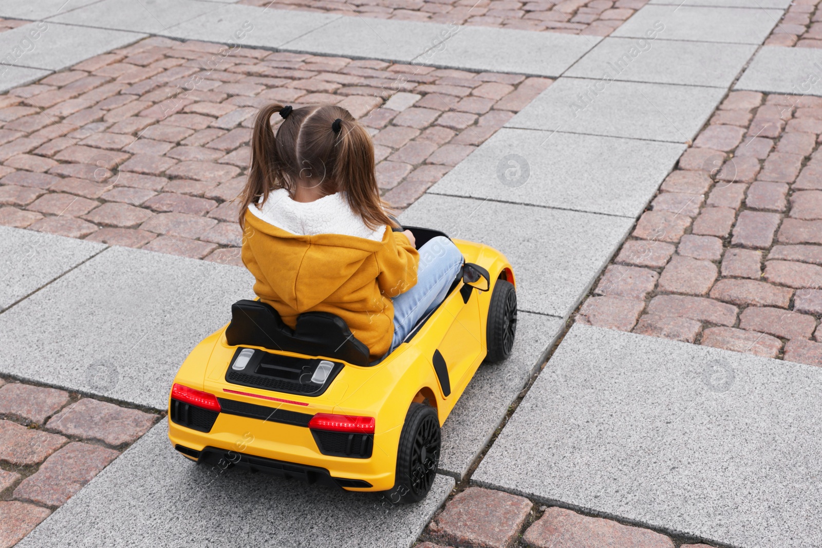 Photo of Cute little girl driving children's car on city street, back view. Space for text