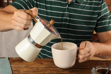 Man pouring aromatic coffee from moka pot into cup at wooden table, closeup