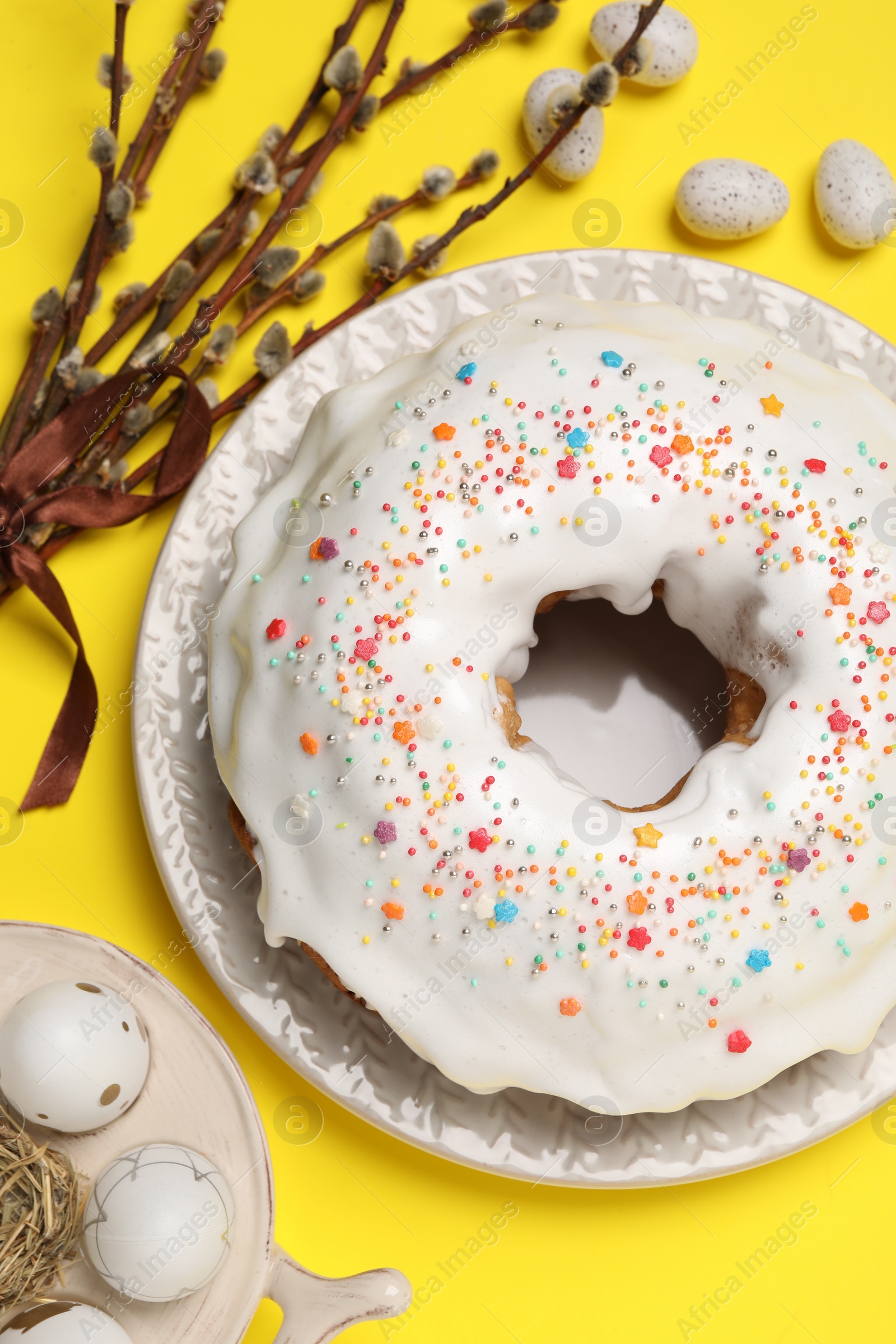 Photo of Easter cake with sprinkles, painted eggs and willow branches on yellow background, flat lay