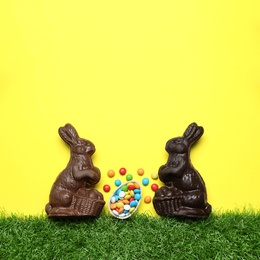 Chocolate Easter bunnies, halved egg and candies with green grass on yellow background