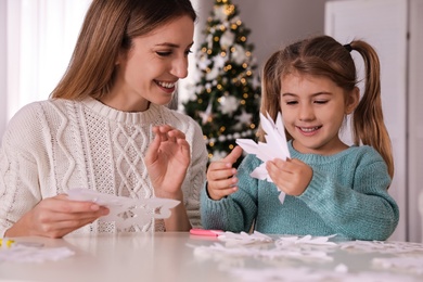 Photo of Happy mother and daughter making paper snowflakes at table near Christmas tree indoors