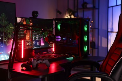 Photo of Playing video games. Stylish room interior with modern computer, wireless controllers and gaming chair in neon lights