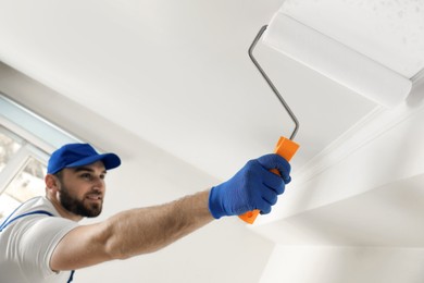 Photo of Handyman painting ceiling with white dye indoors, focus on roller