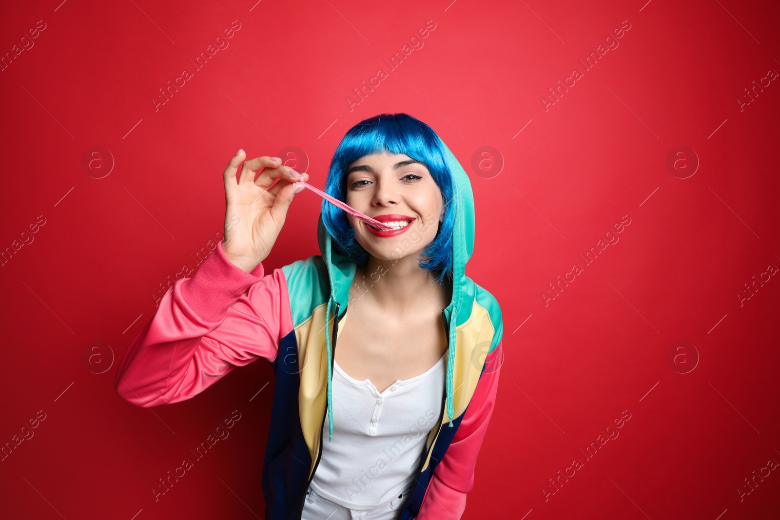 Photo of Fashionable young woman in colorful wig chewing bubblegum on red background