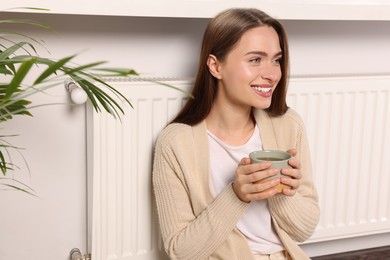 Photo of Woman holding cup with hot drink near heating radiator indoors