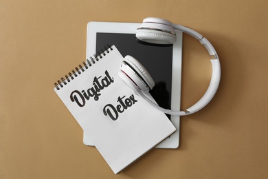 Photo of Tablet, headphones and notebook with words DIGITAL DETOX on beige background, top view