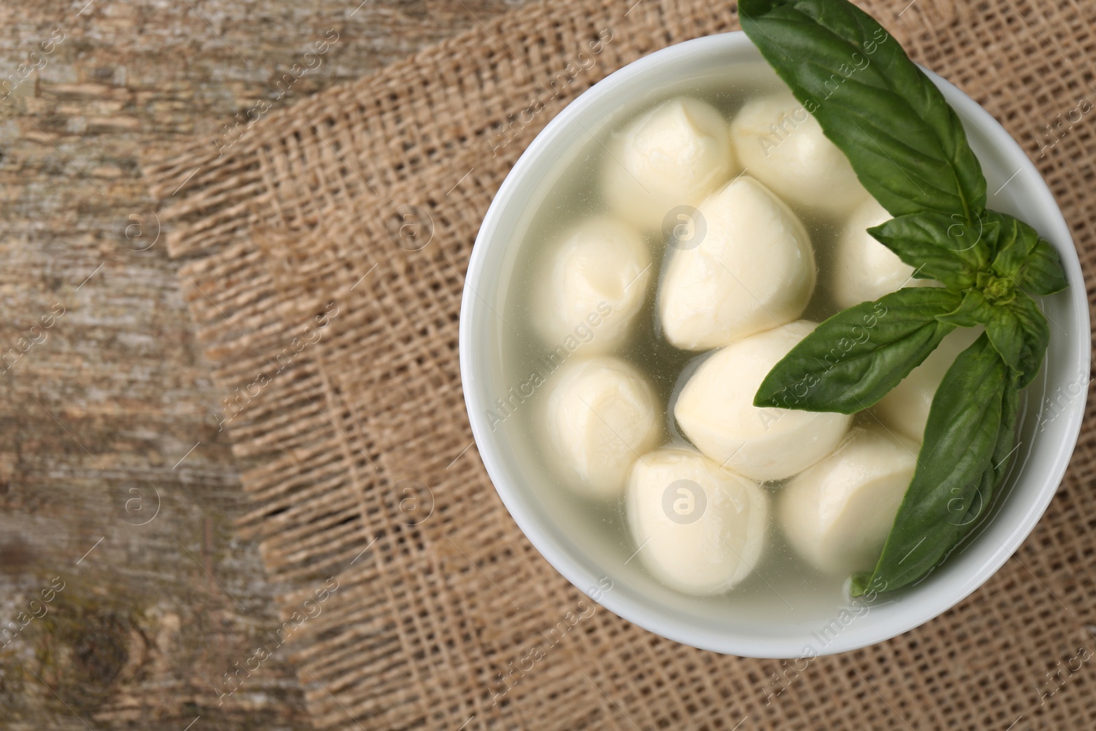 Photo of Tasty mozzarella balls and basil leaves in bowl on wooden table, top view. Space for text