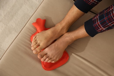 Photo of Man warming feet with hot water bottle on sofa, closeup