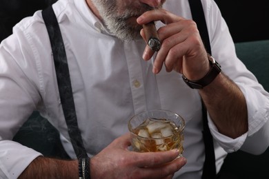 Man with glass of whiskey smoking cigar against black background, closeup