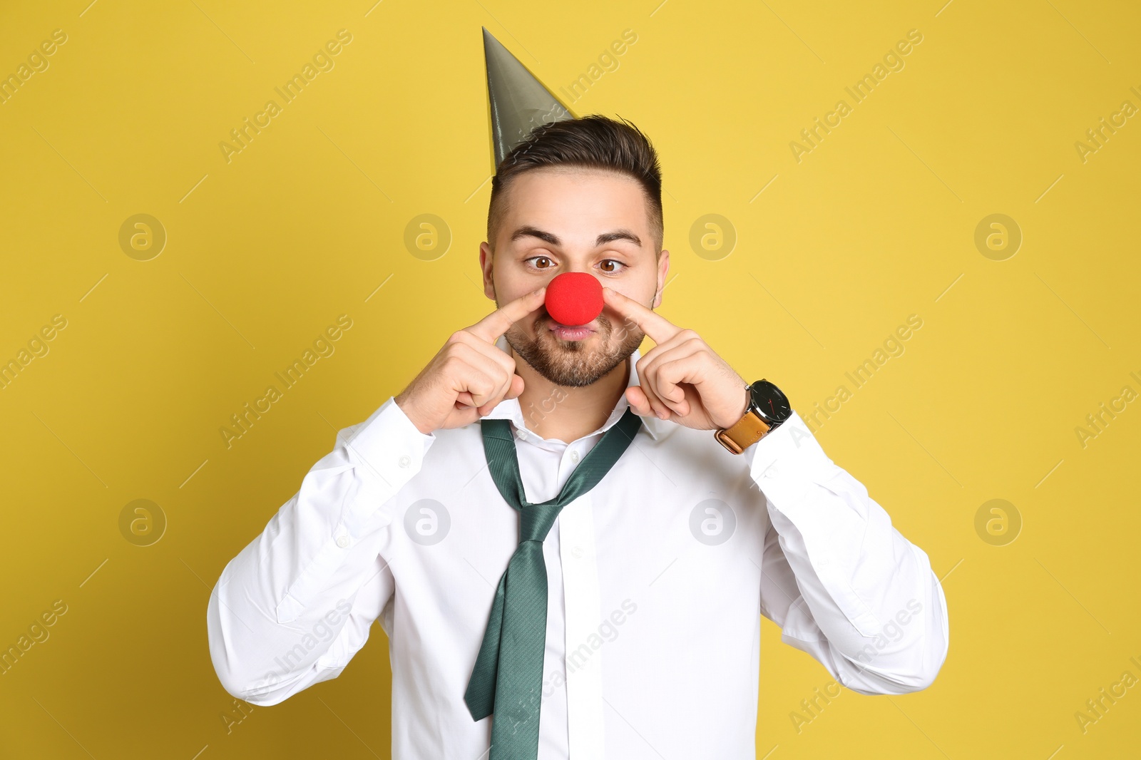 Photo of Emotional young man with party cap and clown nose on yellow background. April fool's day