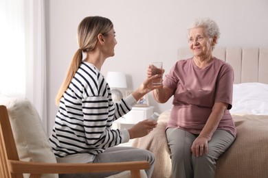 Photo of Young caregiver giving water to senior woman in bedroom. Home care service
