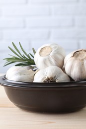 Fresh garlic bulbs in bowl and rosemary on wooden table, closeup