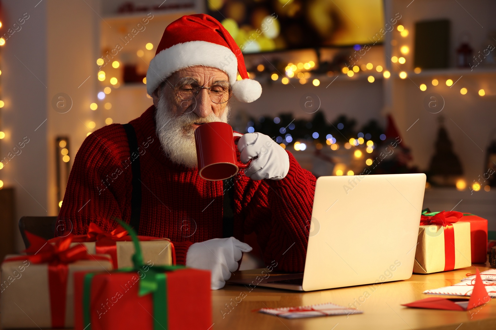 Photo of Santa Claus using laptop and drinking hot beverage at his workplace in room decorated for Christmas