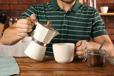 Man pouring aromatic coffee from moka pot into cup at wooden table indoors, closeup