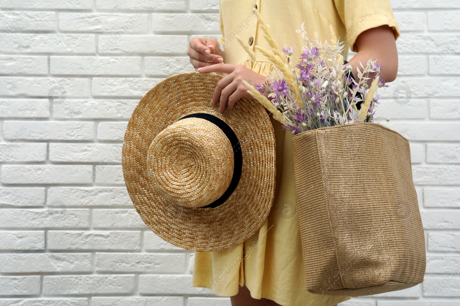 Photo of Woman holding straw hat and beach bag with beautiful bouquet of wildflowers near white brick wall, closeup