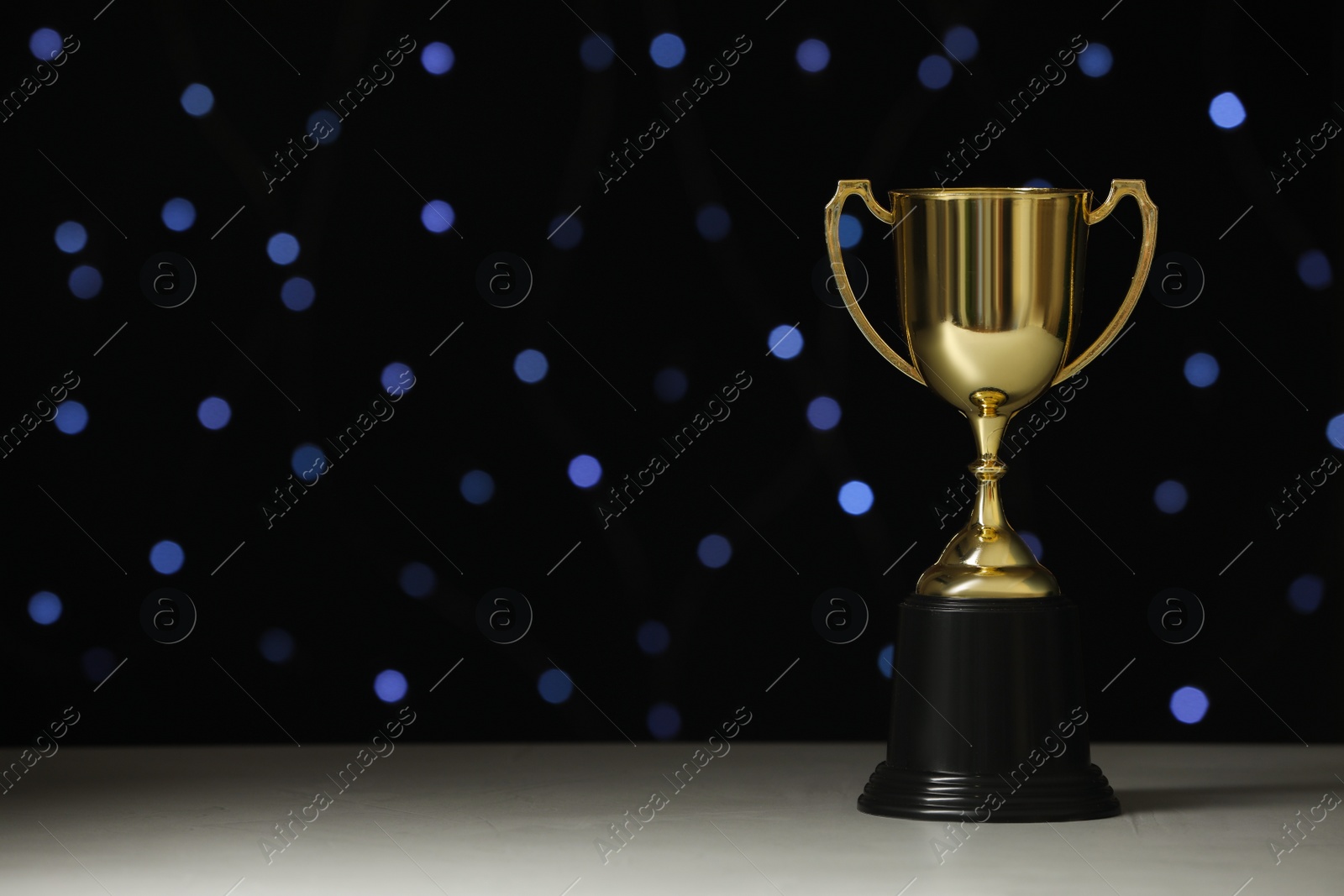Photo of Golden trophy cup on white table against blurred festive lights, space for text