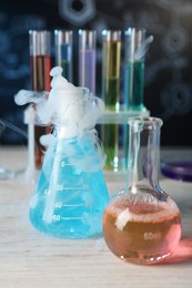 Photo of Laboratory glassware with colorful liquids and steam on white wooden table against black background, closeup. Chemical reaction