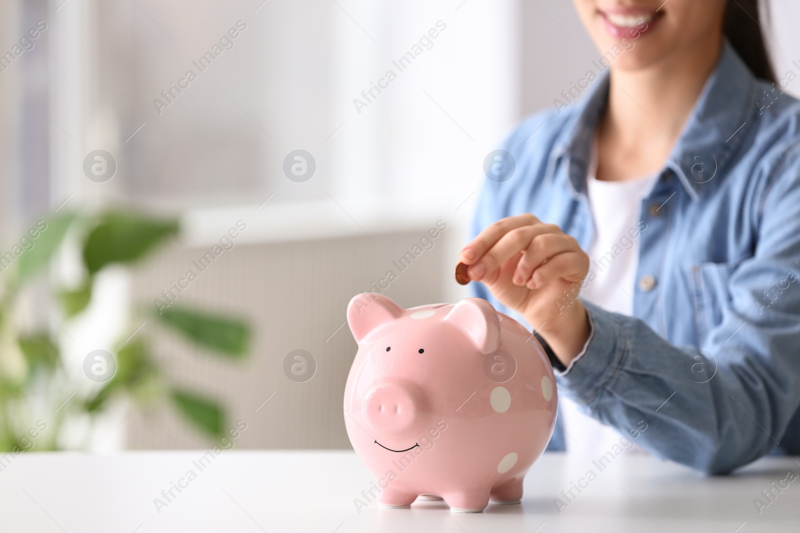 Photo of Woman putting coin into piggy bank at table indoors, closeup. Space for text